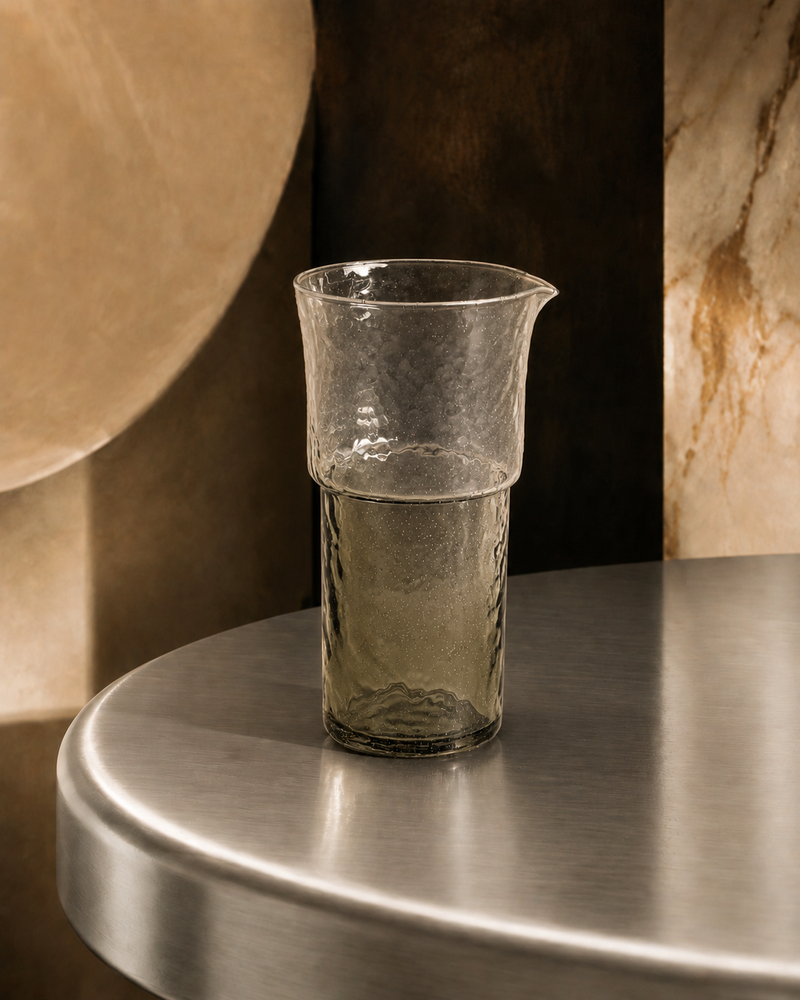 Tall hand-blown textured glass carafe with pouring spout and smoky green-grey gradient base, styled on a metal tabletop