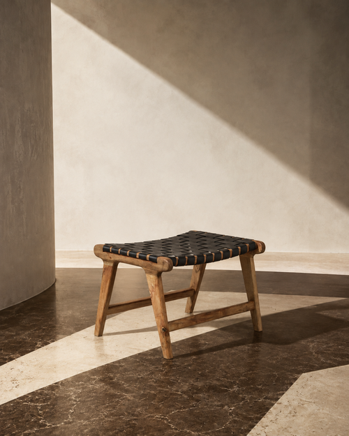 Black woven buffalo leather footstool with natural acacia wood A-frame legs, styled in a sunlit interior